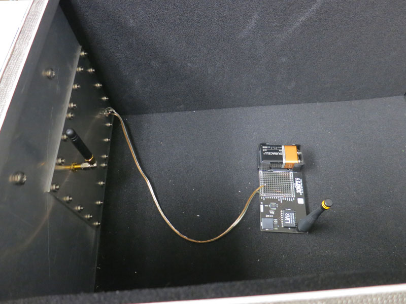 Interior of a foam-lined test enclosure with a small 916 MHz transmitter board and battery on the floor, its antenna a few inches from a second antenna fed through the I/O plate