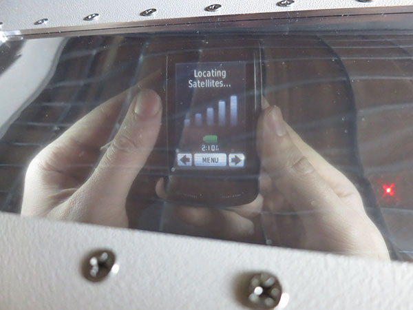 Close-up through the shielded window of a test enclosure: a handheld device is held inside the chamber and its screen, reading Locating Satellites, is sharply legible through the window